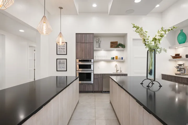 Kitchen featuring dual islands with dark countertops, built-in double ovens, a wet bar with open shelving, recessed lighting, and pendant light fixtures