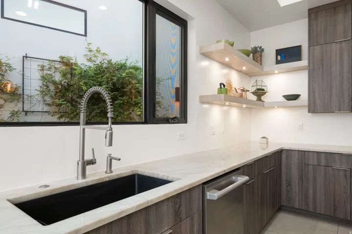 Stainless steel commercial-style faucet over an undermount sink, light-toned countertop, wood-finish cabinetry, floating shelves with integrated lighting, and a built-in dishwasher