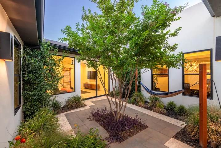 Courtyard featuring a central tree, paved pathways, lush landscaping, exterior wall lighting, and large windows