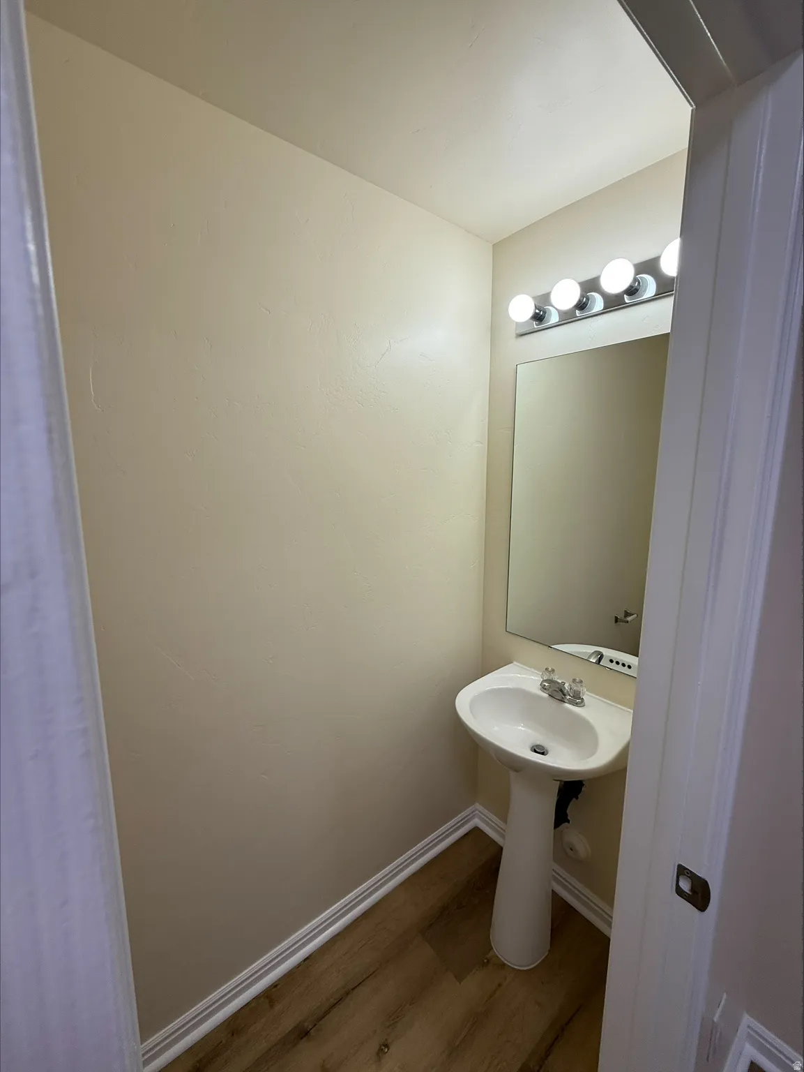 Pedestal sink with chrome fixtures, mirror, vanity lighting, and wood-finish flooring