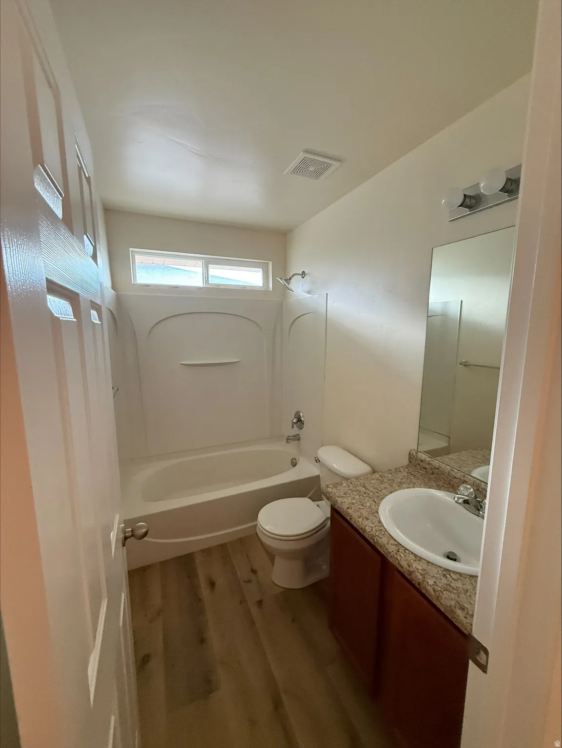 White bathtub and shower combination with a single-handle faucet, vanity with a light-toned countertop, integrated sink, and wood-finish cabinetry, and wood-finish flooring