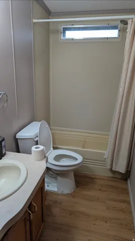 Bathroom featuring a bathtub with an integrated shower, a white toilet, a vanity with a light-colored countertop and integrated sink, wood-finish flooring, and a horizontal transom window
