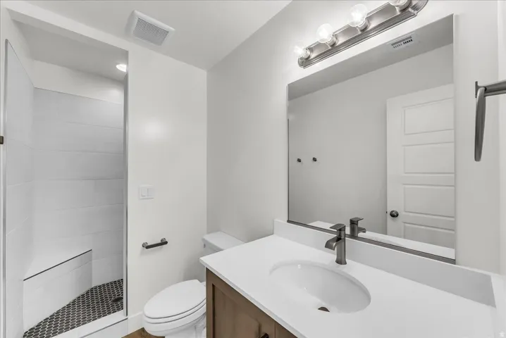 Walk-in shower with tiled walls and hex floor, wood-finish vanity with white countertop, oval undermount sink, and wall-mounted vanity lighting