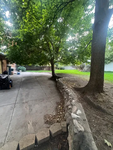 Spacious backyard featuring mature trees, a concrete patio, a stone retaining wall, and green lawn space