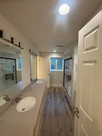 Dual vanity with integrated sinks, wood-finish flooring, recessed lighting, skylight, and enclosed shower with glass doors