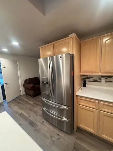 Wood-finish flooring with a stainless steel refrigerator, light wood cabinetry, and a subway tile backsplash