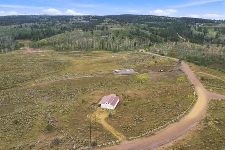 Expansive terrain with a white outbuilding featuring a red roof, bordered by a dirt road, and surrounded by varied natural landscaping