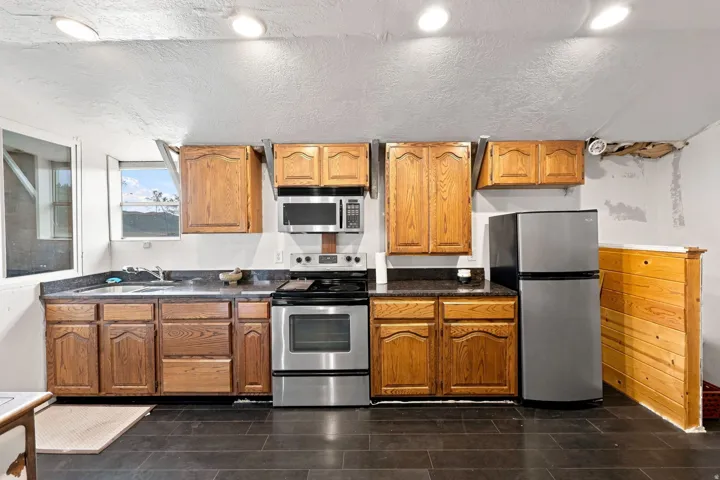 Kitchen area featuring wood-finish cabinetry, stainless steel appliances, dark countertops, and dark tile flooring