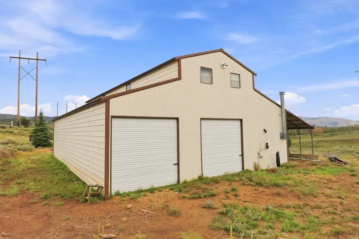 Metal-sided barn structure with a gable roof, featuring two roll-up garage doors, a covered lean-to extension, and two upper windows