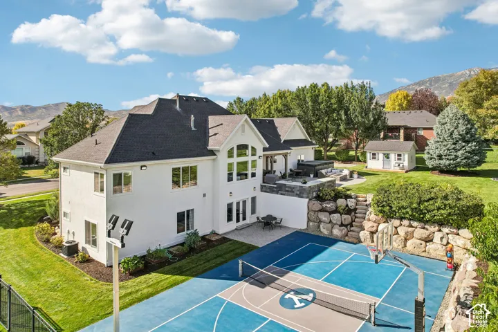 Stucco exterior with multiple windows, elevated patio with a built-in spa, terraced landscaping with rock retaining walls, sport court, and a detached shed