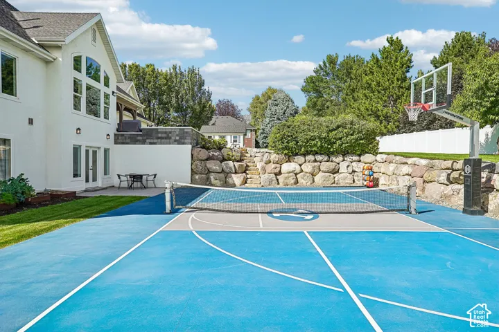 Multipurpose sports court with basketball hoop and tennis net, expansive stucco exterior, arched window array, upper-level balcony, and stone retaining wall