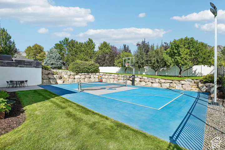 Multi-sport court with basketball hoop and net, surrounded by a stone retaining wall, mature trees, and a white fence