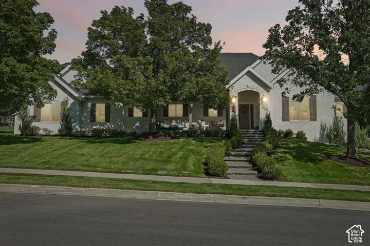Stucco exterior with a peaked roofline, manicured lawn, stone pathway, and mature trees