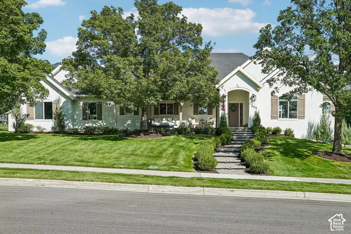 Stucco exterior with wood shutters, front porch with stone steps, mature trees, and landscaped lawn