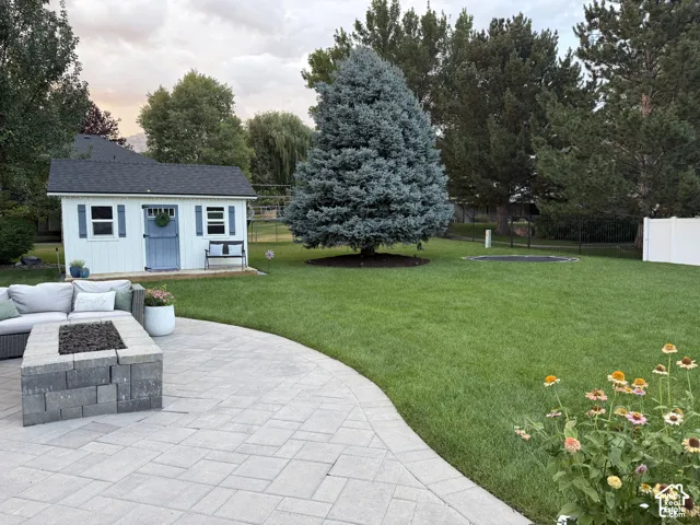 Expansive backyard featuring a large paved patio, a built-in fire pit, a storage shed with blue shutters, mature trees, and a manicured lawn