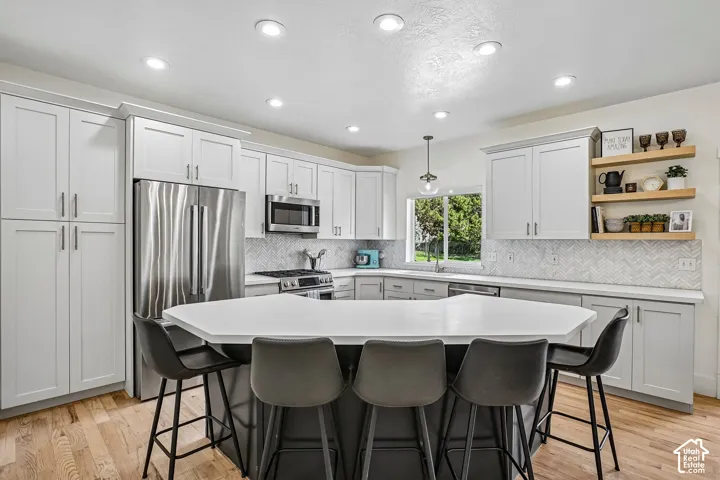 Kitchen featuring light-finish cabinetry, stainless steel appliances, a chevron-patterned tile backsplash, a large central island with a solid surface countertop, and wood-finish flooring