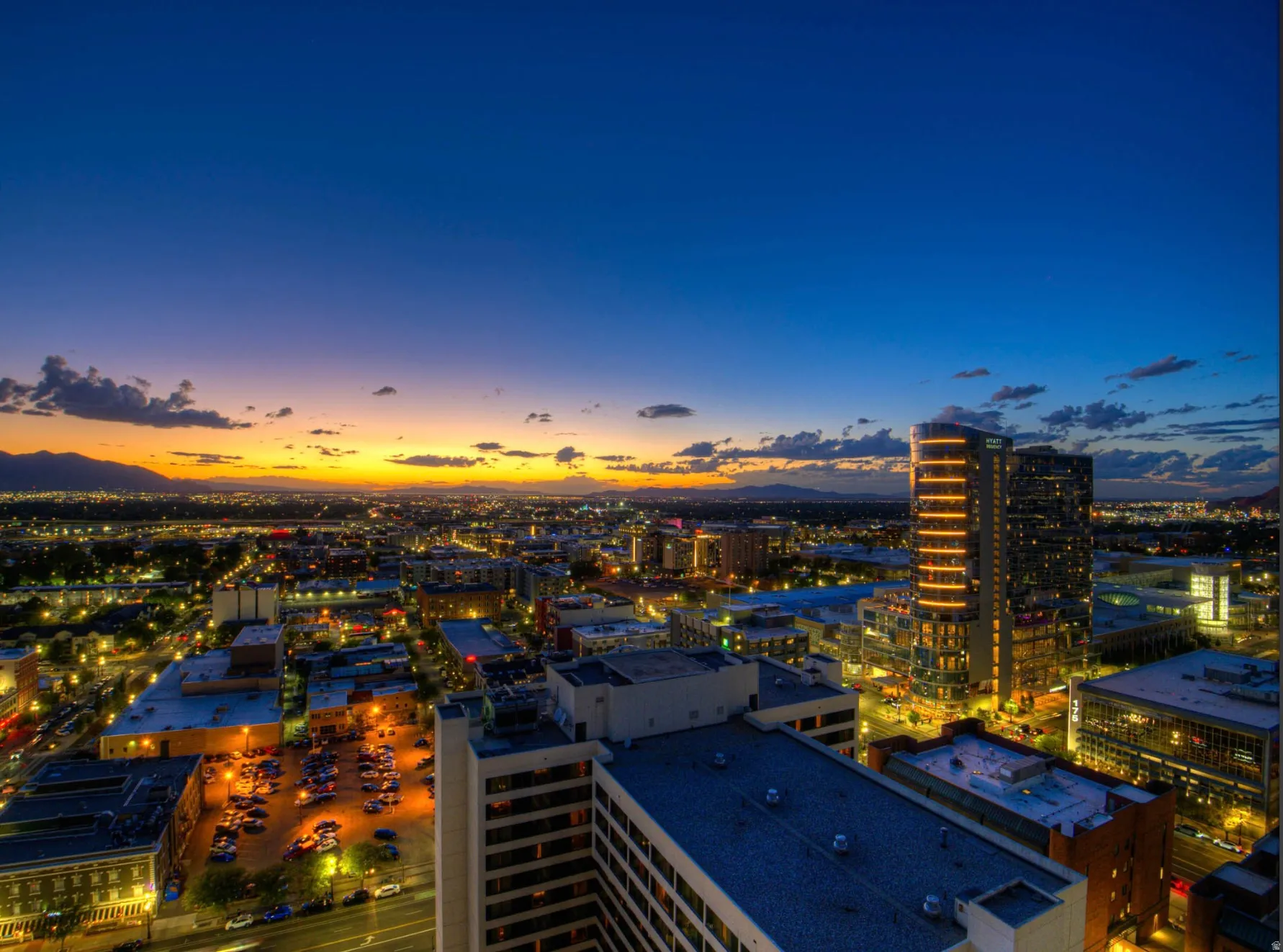 Panoramic cityscape with distant mountains, illuminated buildings, and a vibrant sunset horizon
