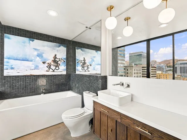Freestanding rectangular bathtub, dark tile accent wall, wood-finish vanity with rectangular vessel sink, white countertop, and full-height window with urban scenery
