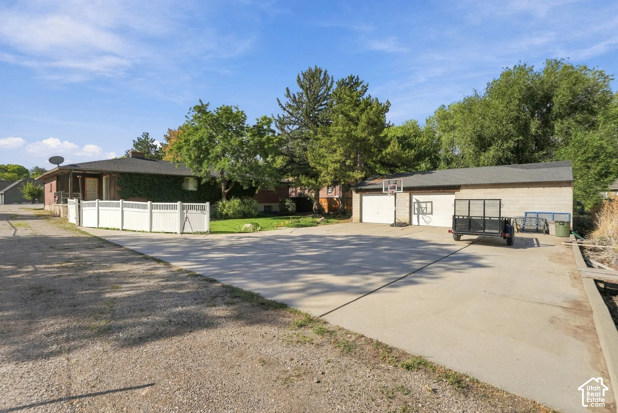Expansive concrete paving, two-car garage with white doors, surrounding mature trees, and a low white fence
