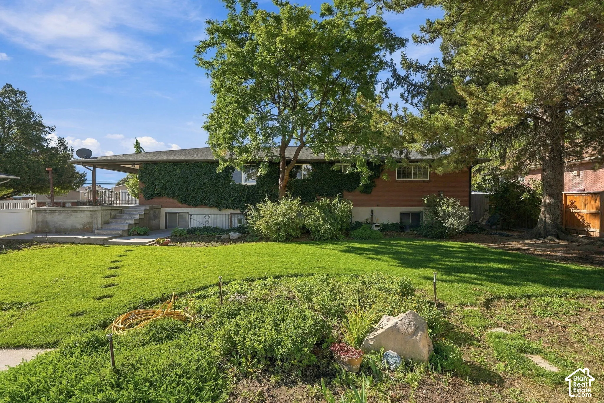 Brick exterior with extensive green climbing foliage, mature trees, a covered patio area, and a terraced lawn with stepping stones