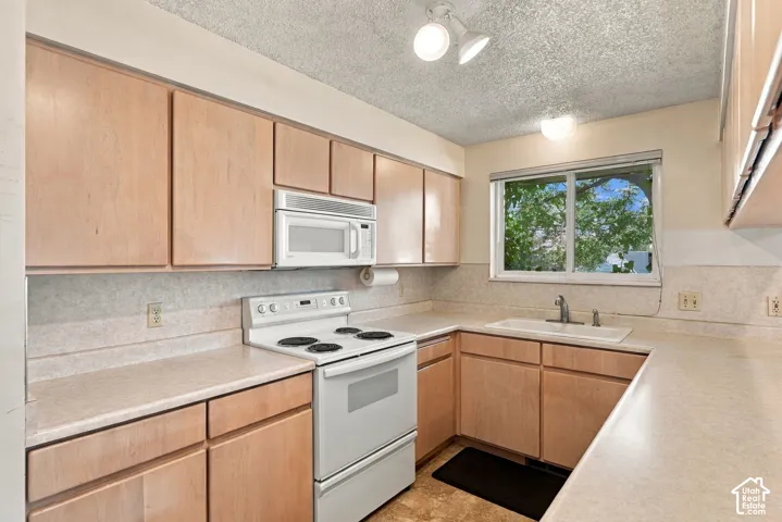 Galley kitchen featuring warm wood-finish cabinetry, white electric range, stainless steel microwave, tile backsplash, and light-colored countertops
