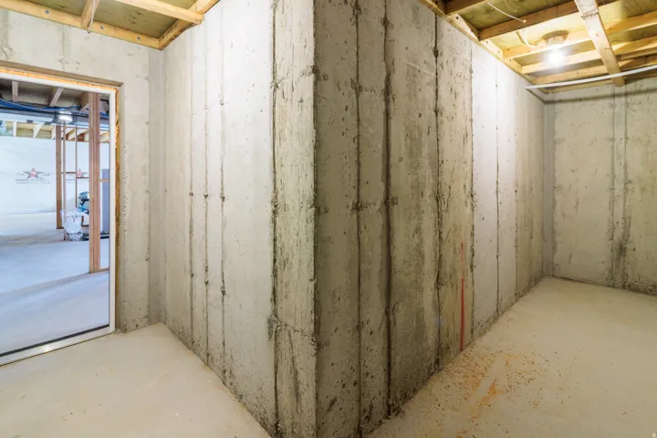 Unfinished basement featuring concrete walls and flooring, exposed ceiling joists, and a doorway with a wood frame