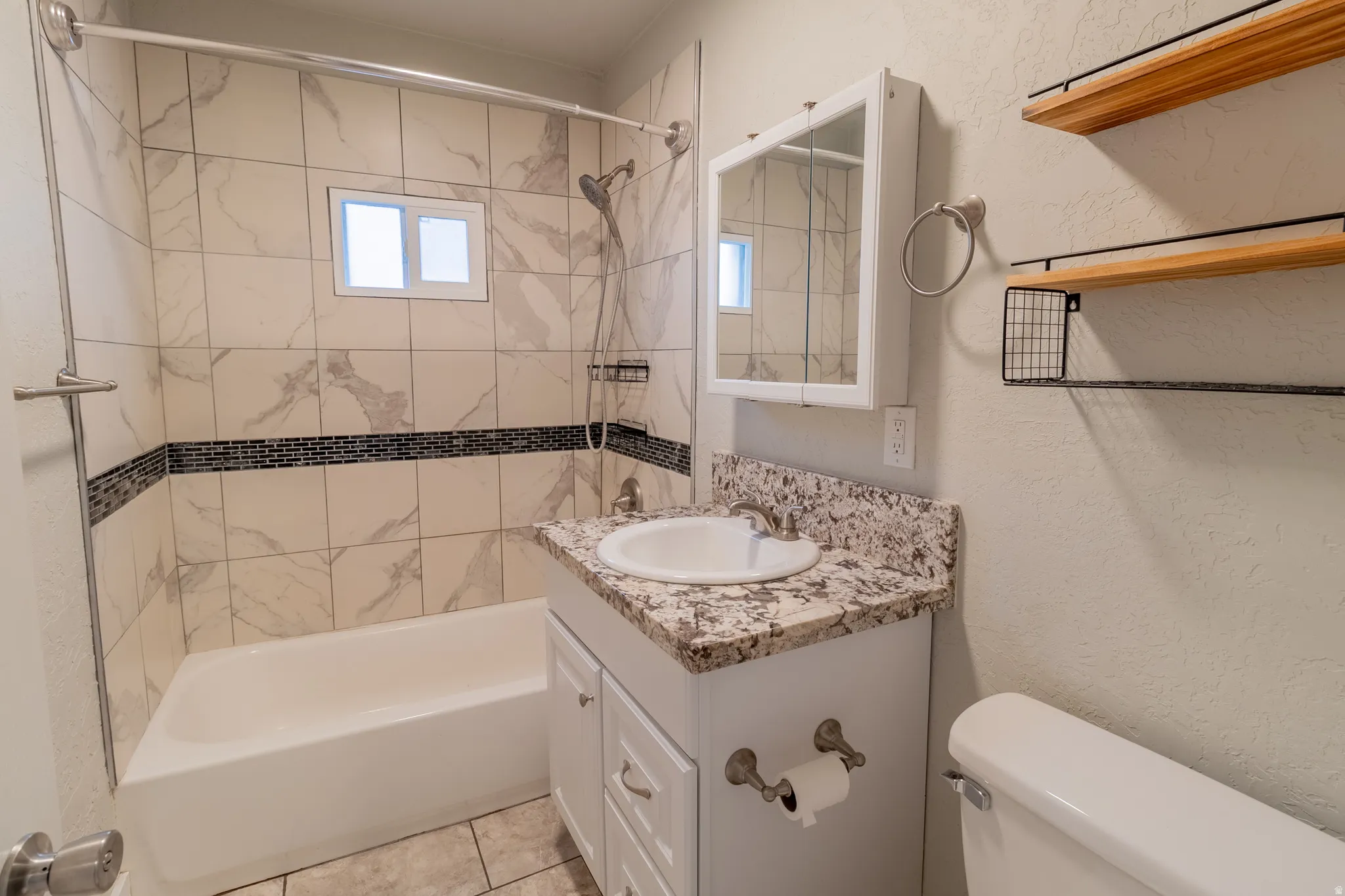 Bathtub with tiled surround featuring a mosaic accent band, vanity with a stone-finish countertop, and wall-mounted shelving with metal accents