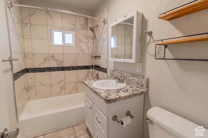 Bathtub with tiled surround featuring a mosaic accent band, vanity with a stone-finish countertop, and wall-mounted shelving with metal accents