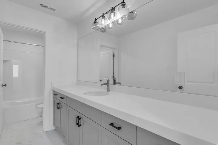 Full secondary bath with extra-long vanity, painted wood cabinets, quartz slab countertops and door between tub and sink.