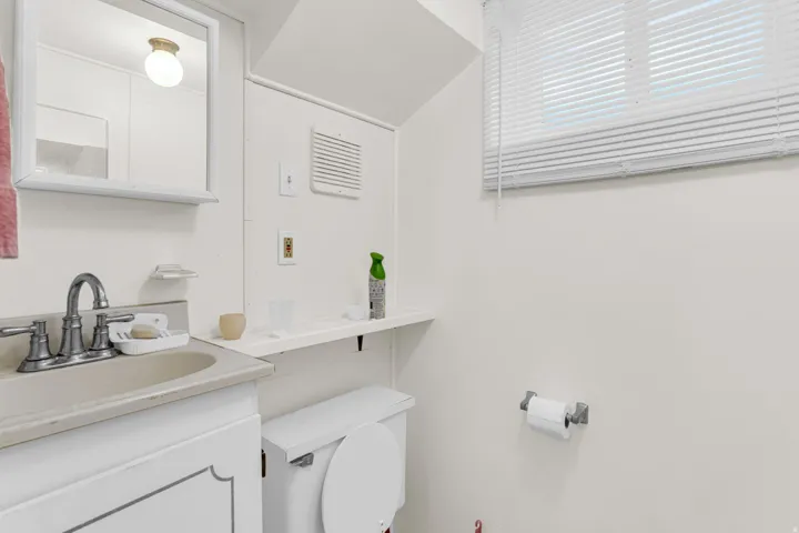 Vanity with integrated sink, wall-mounted mirror, window with blinds, toilet, and flush-mount ceiling light