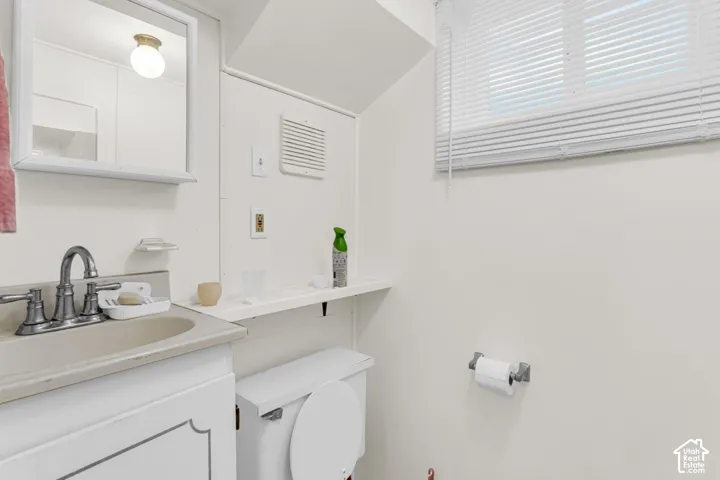 Vanity with integrated sink, wall-mounted mirror, window with blinds, toilet, and flush-mount ceiling light