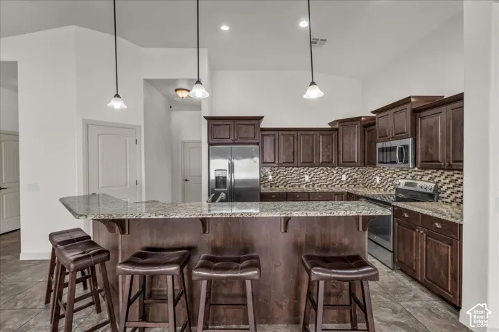 Kitchen featuring dark wood cabinetry, stainless steel appliances, a breakfast bar with granite-style countertop, and a mosaic tile backsplash