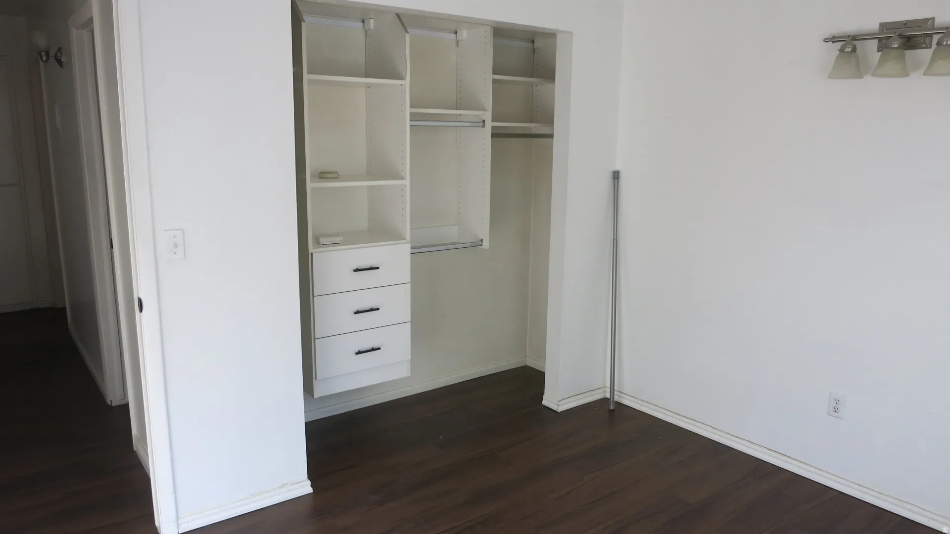 Bedroom 1 - Wood-finish flooring with white baseboards, built-in closet system with shelving and drawers, and a wall-mounted light fixture with three sconces