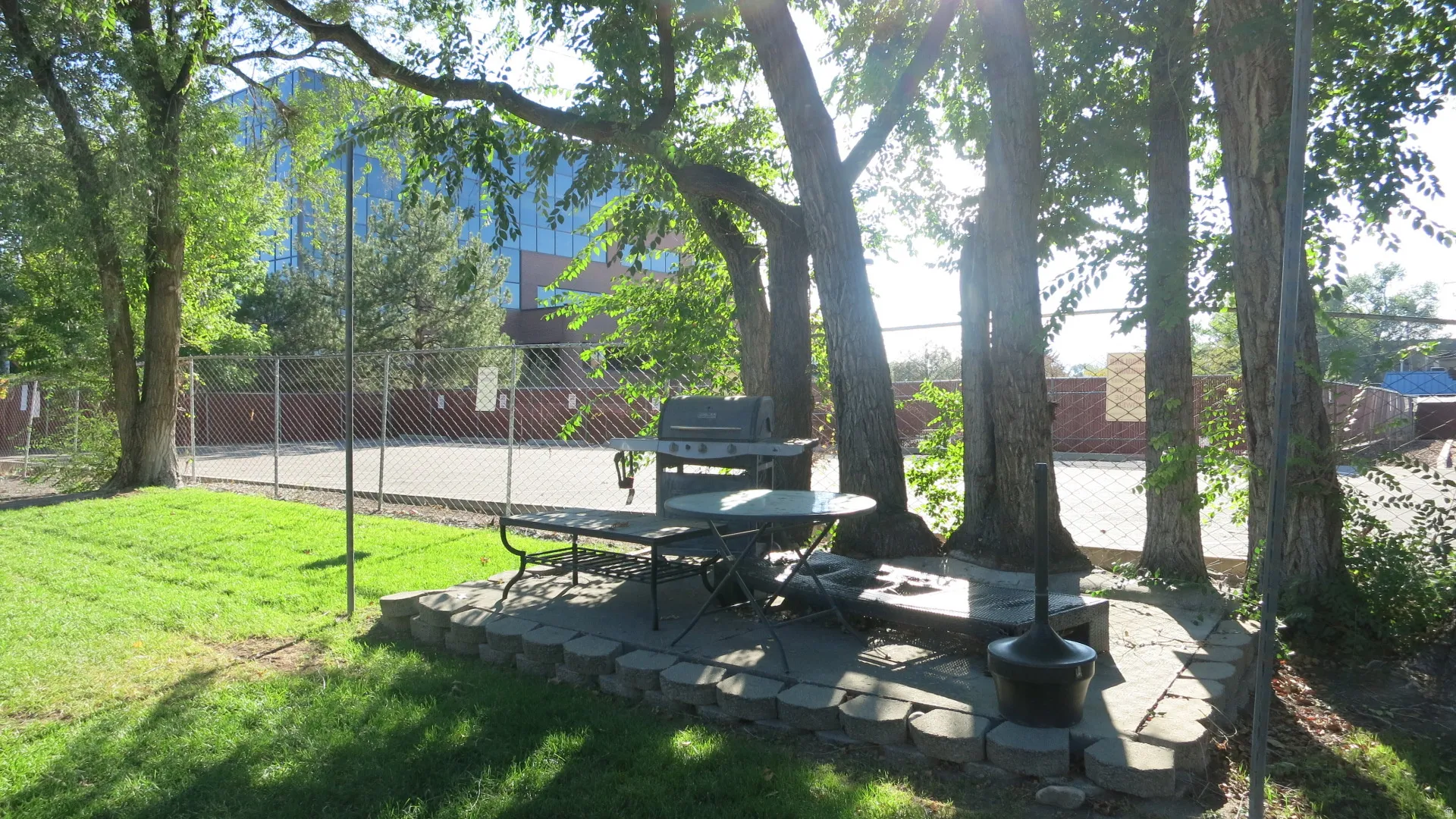 Expansive lawn area with mature trees, a paver patio, barbeque and a chain-link fence
