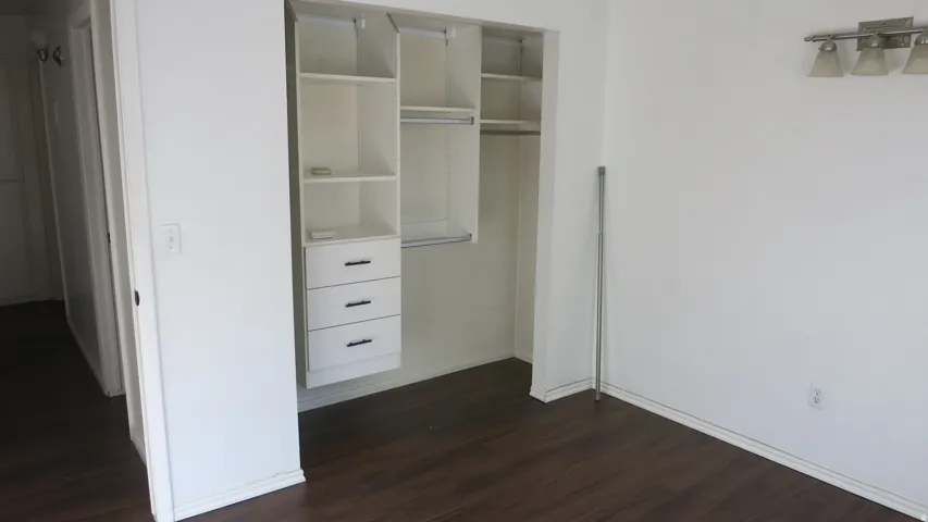Bedroom 1 - Wood-finish flooring with white baseboards, built-in closet system with shelving and drawers, and a wall-mounted light fixture with three sconces