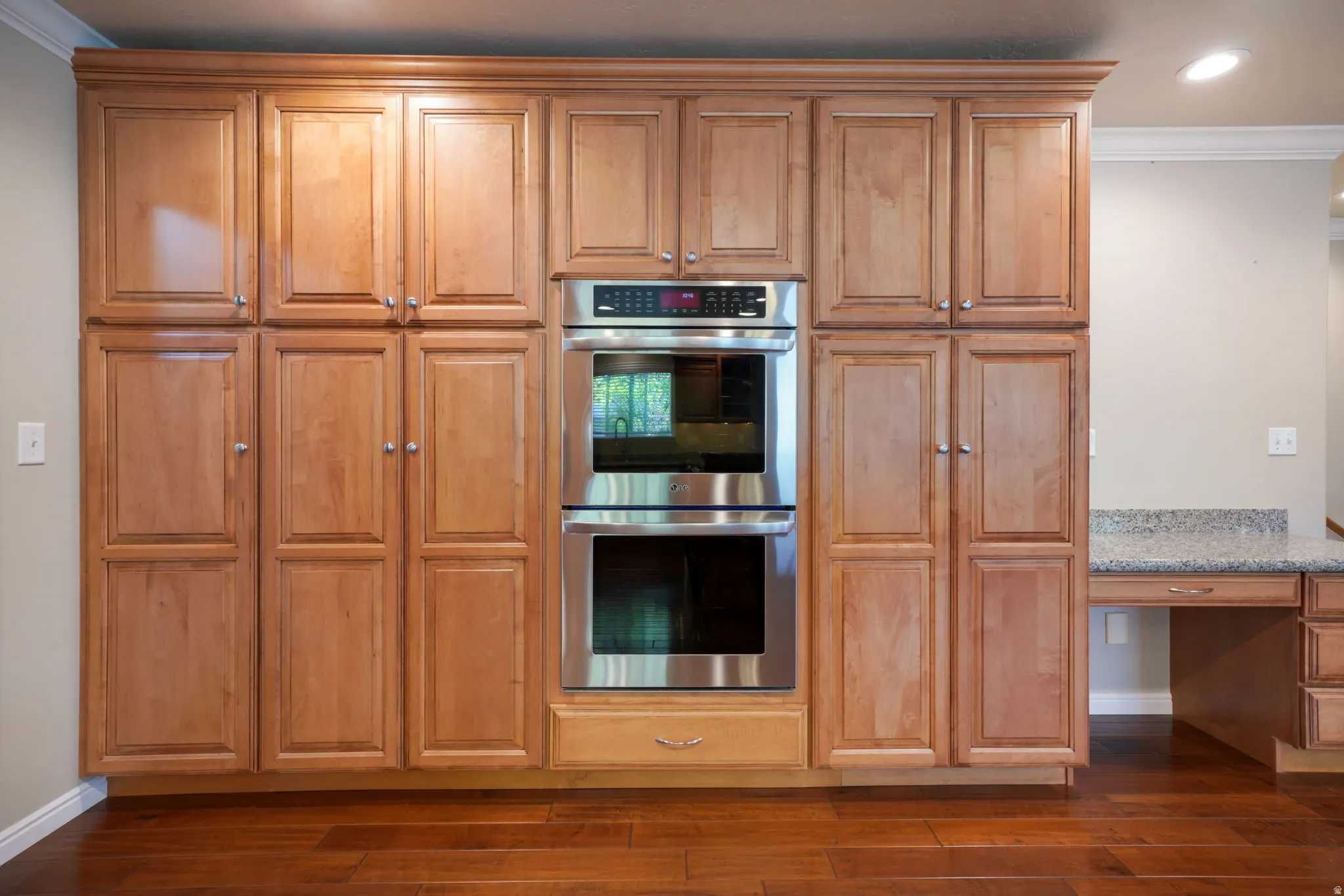 Stacked wood cabinetry with crown molding, stainless steel double wall ovens, wood-finish flooring, and a built-in desk with a stone countertop