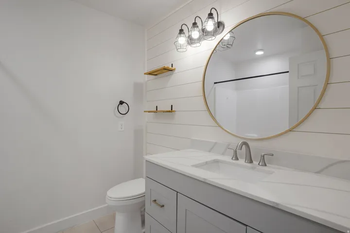 White shiplap accent wall, light grey vanity with integrated sink, white countertop with veining, round gold-framed mirror, and industrial-style vanity lighting