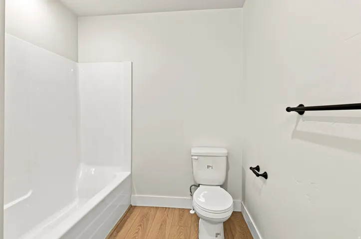 White bathtub and shower combination unit, white toilet, wood-finish flooring, and matte black towel bar and toilet paper holder