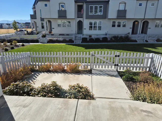 White picket fence with gate, concrete walkway, landscaped garden beds, and manicured lawn