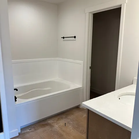 White bathtub with matte black fixtures, wainscoting-style wall surround, wood-finish flooring, and a vanity with a light-toned countertop