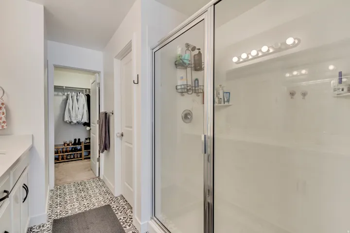 Glass-enclosed shower, built-in shower shelving, vanity with white countertop, patterned floor tile, and a walk-in closet with built-in storage