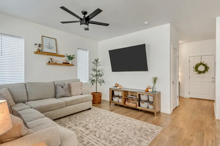 Light wood-finish flooring, white walls, multiple windows with blinds, a black ceiling fan, and a front entry door