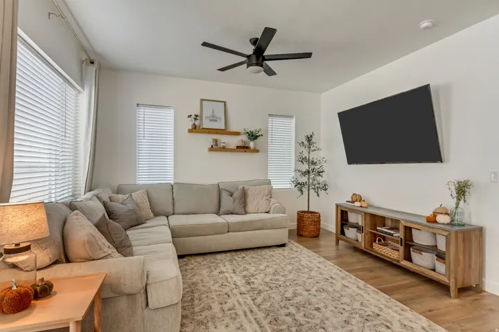 Wood-finish flooring throughout, ceiling fan with integrated lighting, multiple windows with blinds, wall-mounted television, and floating wood shelving