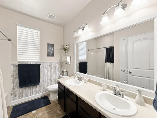 Dual vanity with integrated sinks and chrome fixtures, framed mirror, wall-mounted light fixture, wood-finish wainscoting, and ceramic tile flooring