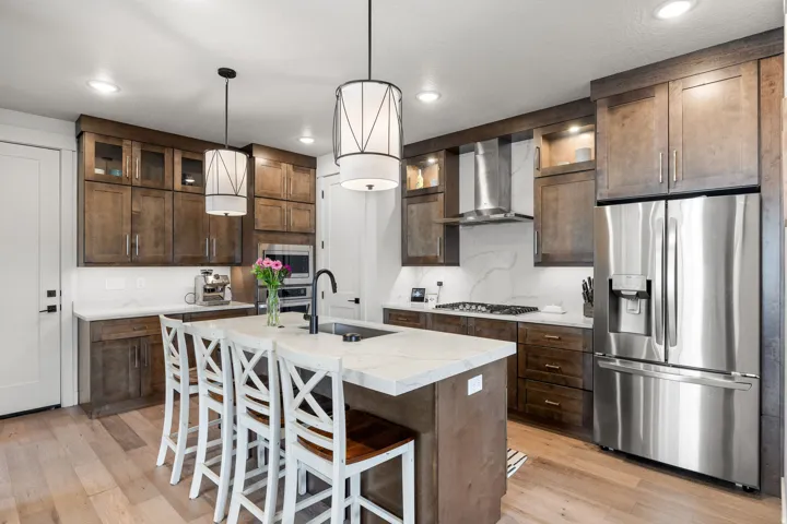 Hardwood flooring, dark wood-finish cabinetry, stainless steel appliances, white countertop island with integrated sink, and recessed lighting with pendant fixtures