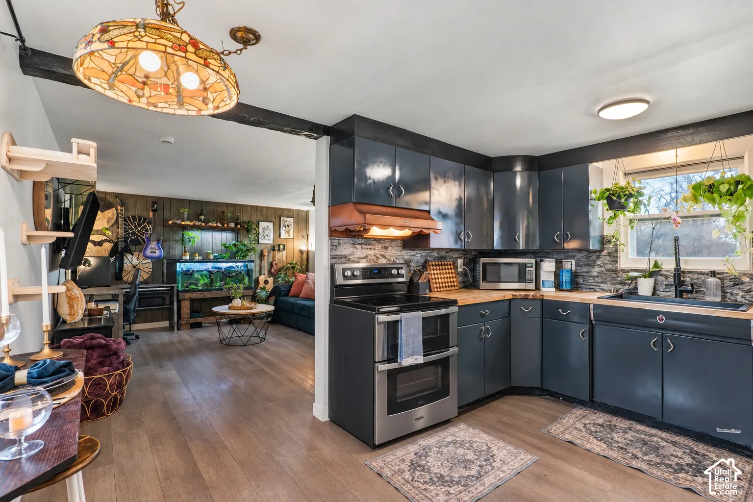 Original photo shows the old stove, this was altered to show the new stove.Kitchen featuring dark cabinetry, stainless steel appliances, wood-finish countertops, and a stacked stone-look backsplash