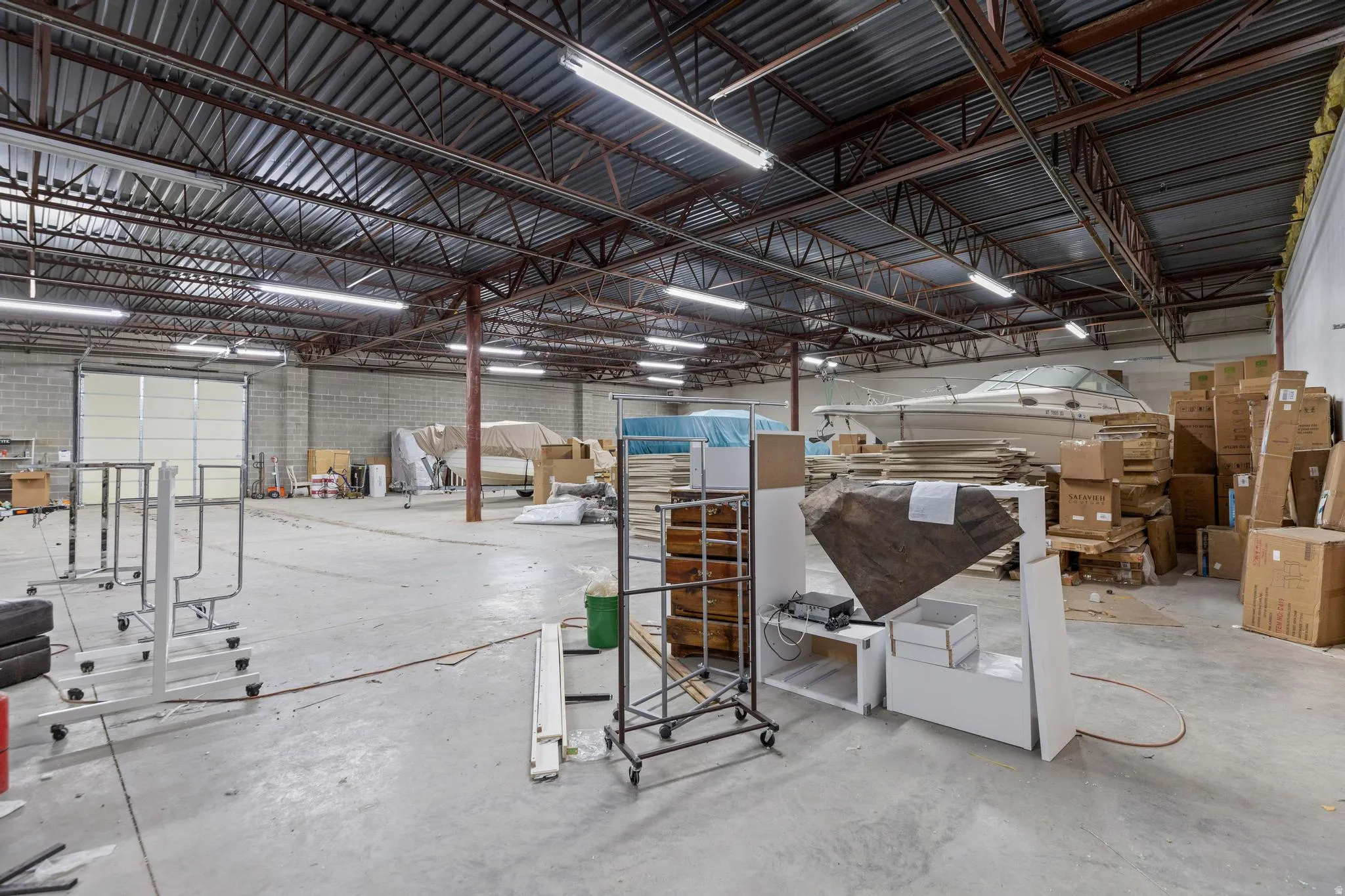 Expansive warehouse space featuring a high corrugated ceiling, exposed steel trusses, concrete block walls, and overhead fluorescent lighting