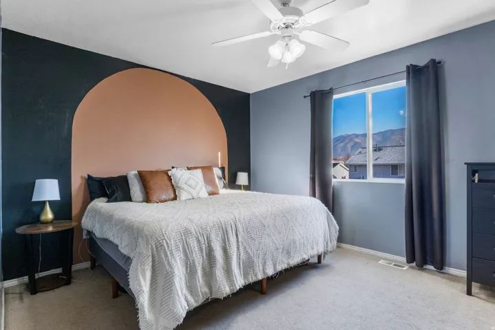 Carpeted flooring, ceiling fan with integrated lighting, wall-mounted window with dark drapery, and a distinctive accent wall with an arched design