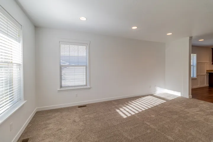 Recessed ceiling lighting illuminates a room with light-colored carpeting, multiple windows, and white window blinds