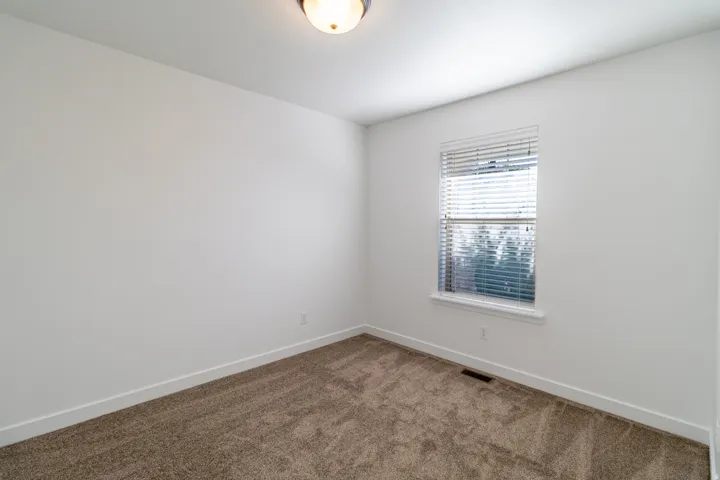 Neutral wall color with plush carpeting, featuring a window with blinds and a ceiling-mounted light fixture